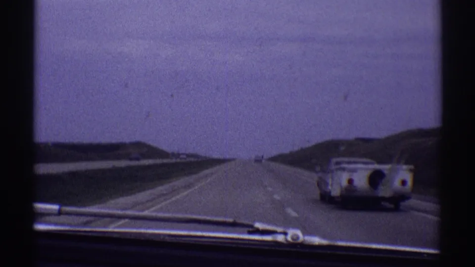 Late-1960s 8mm road view from inside a moving car, with a camper-towing vehicle near the right edge and distant highway traffic.