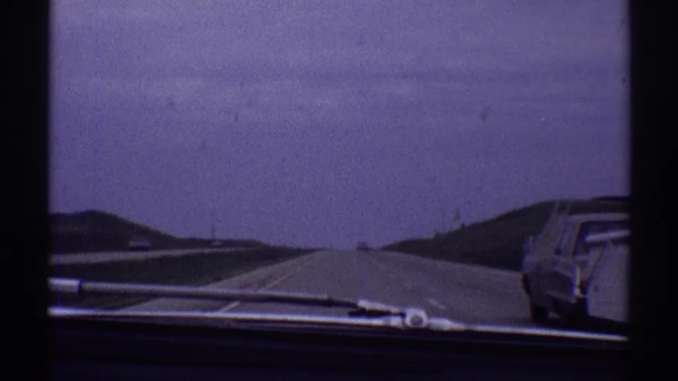 1969 8mm view from inside a car on an open highway near Clarendon Hills, with a sedan by the road and a hazy sky ahead.