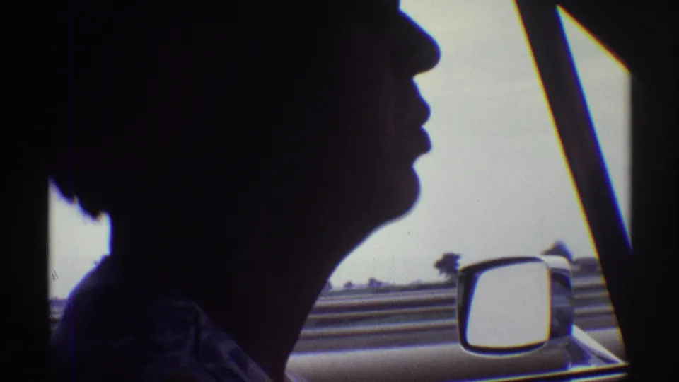 Silhouetted driver seen from the passenger side of a car, with the window frame, side mirror, and highway landscape outside.