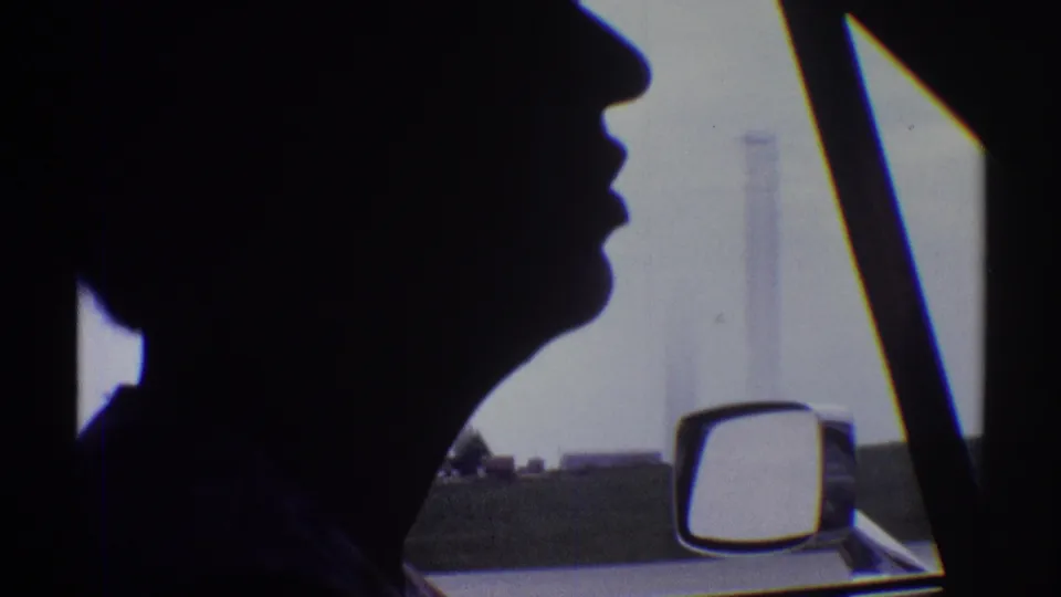 Passenger-side view of the silhouetted driver, with the car mirror, roadside horizon, and faint distant structures visible through the window.