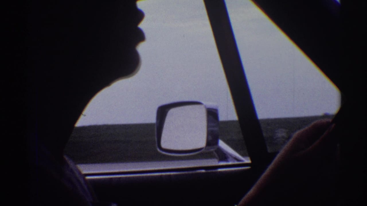 1969 8mm frame of the driver's dark profile against a pale sky, with the car mirror and roadside visible through the window.