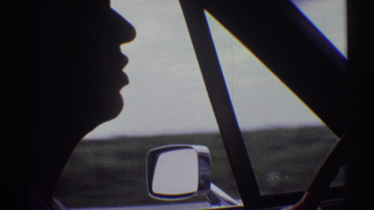 Backlit 1969 home-movie view of a male driver chewing gum, seen from the passenger area with the side mirror in frame.