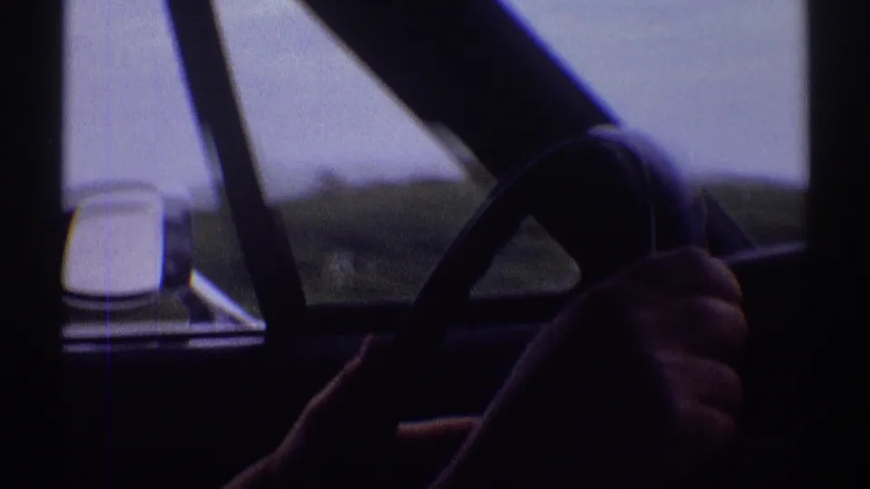 1969 Clarendon Hills frame showing the steering wheel, the driver's hands, side mirror, window frame, and blurred open landscape.