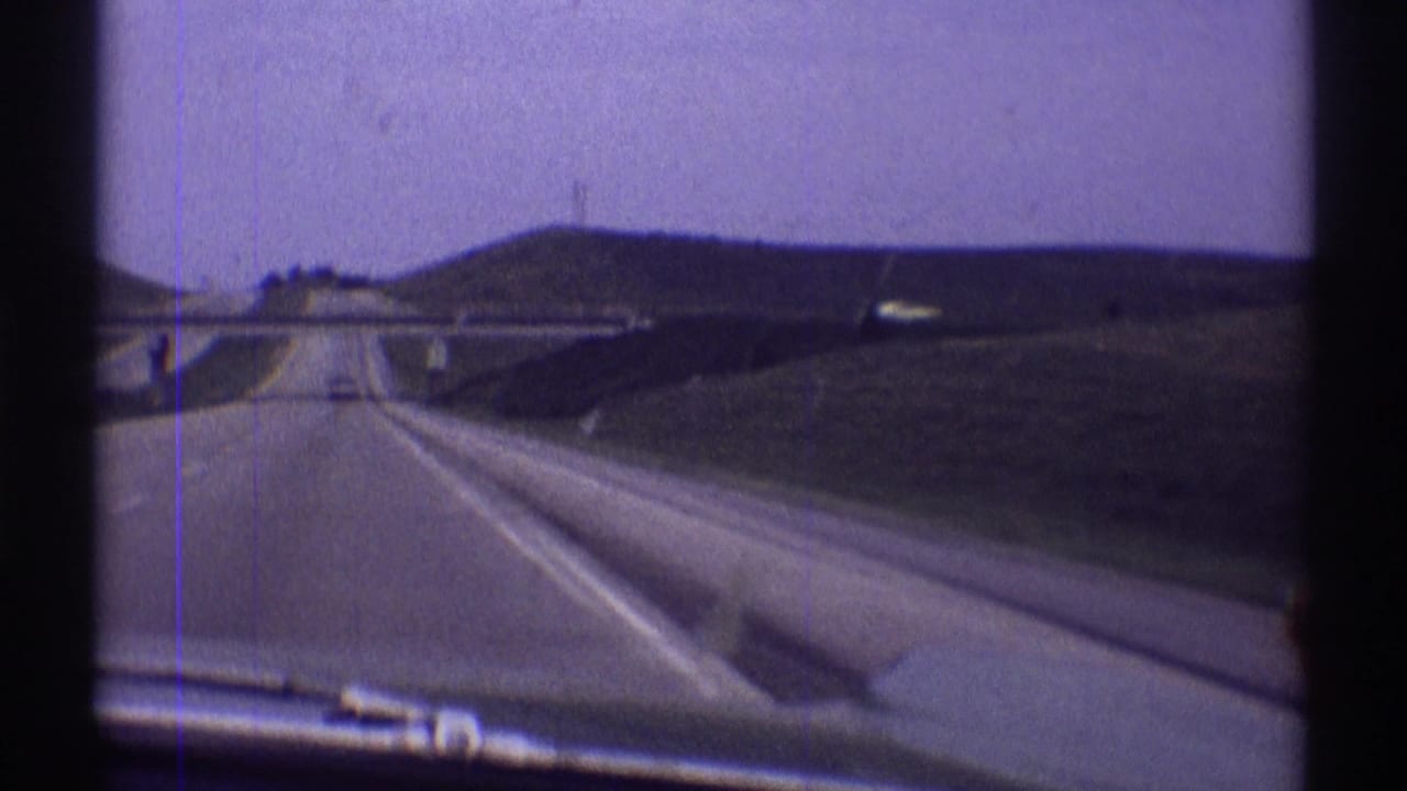 Forward-facing 1969 home-movie view along an open Illinois highway toward an overpass, with lanes, grassy embankments, and pale sky visible.