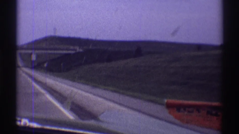 Soft 8mm frame from the moving car showing the highway shoulder, overpass, grassy embankment, and a partial red roadside sign in Clarendon Hills footage.