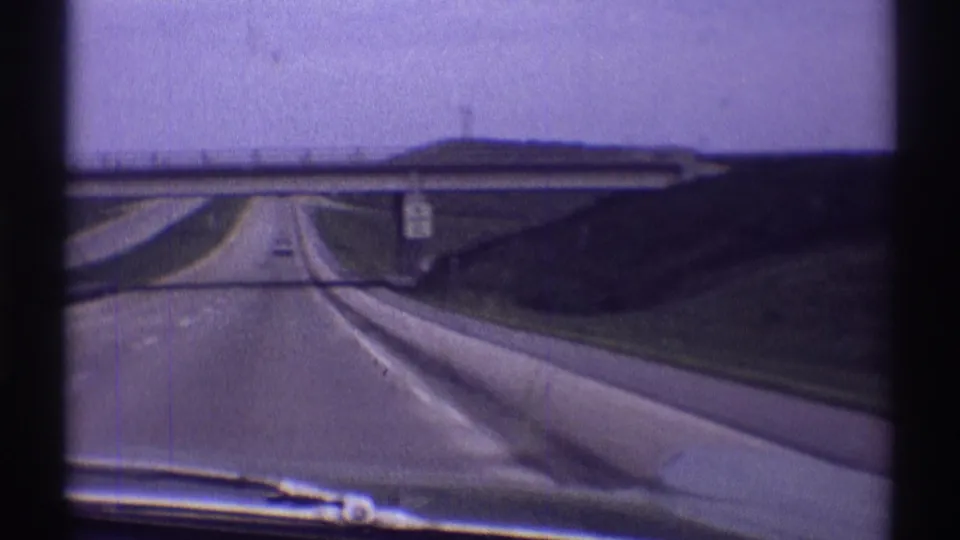 1969 8mm forward-driving view on an open Illinois highway, showing the windshield edge, overpass, grassy banks, lane markings, and a distant car.