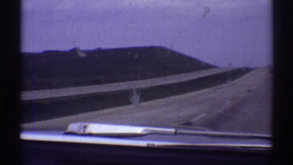 1969 8mm highway POV near Clarendon Hills, Illinois, with the car continuing along a wide road bordered by grassy banks.