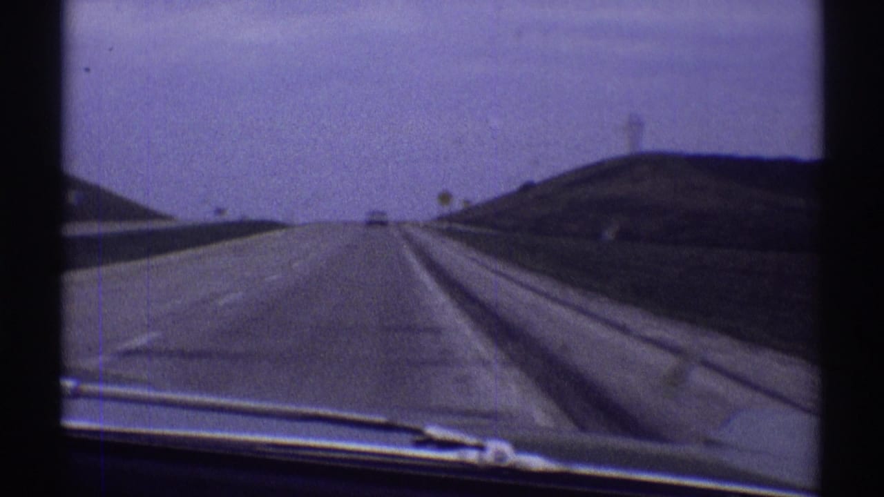 Forward view through a car windshield toward an open road and low roadside hills.