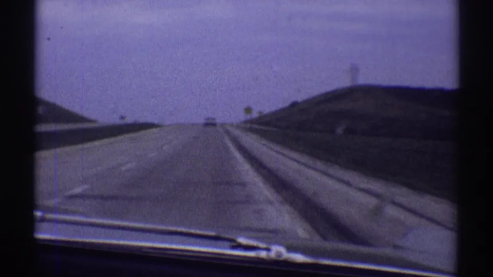 1969 home-movie view from a moving car in Illinois, showing a broad road, windshield edge, wiper, utility poles, and low rural slopes.
