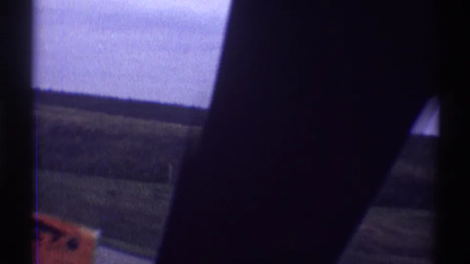 A moving 8mm car-window view in Illinois shows a dark interior pillar, rural field, low horizon, pale sky, and a partly cropped red sign.