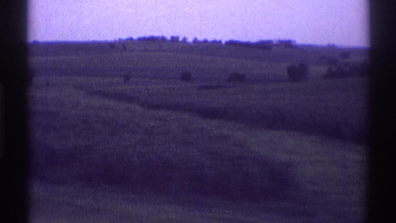 Rural Illinois farmland seen from a moving viewpoint, with open fields, distant tree lines, and dark scan edges from 8mm film.