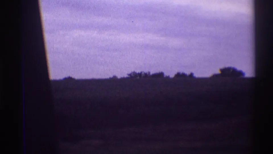Late-1960s 8mm farmland view with a dark foreground, low tree line, cloudy purple sky, and visible film-edge shading.