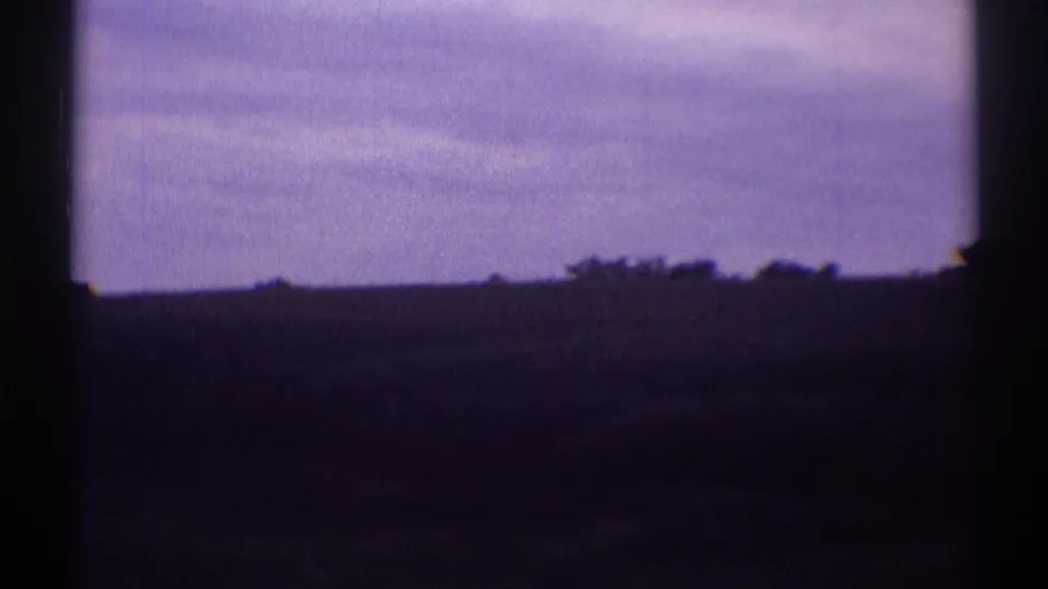 1969 8mm travel footage looking through a dark vehicle window at a flat rural horizon and overcast sky.