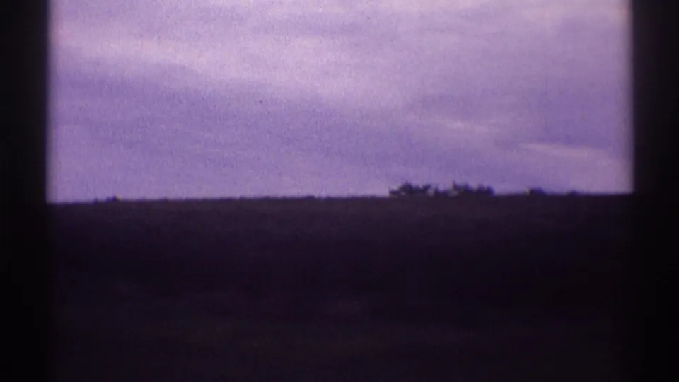 A dark vehicle-window edge frames an overcast 1969 roadside landscape with distant shrubs and a low horizon.