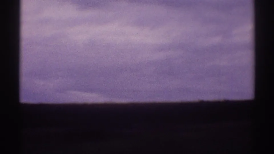 Soft 8mm frame from a moving vehicle, showing a cloudy sky above a dark, low rural horizon.