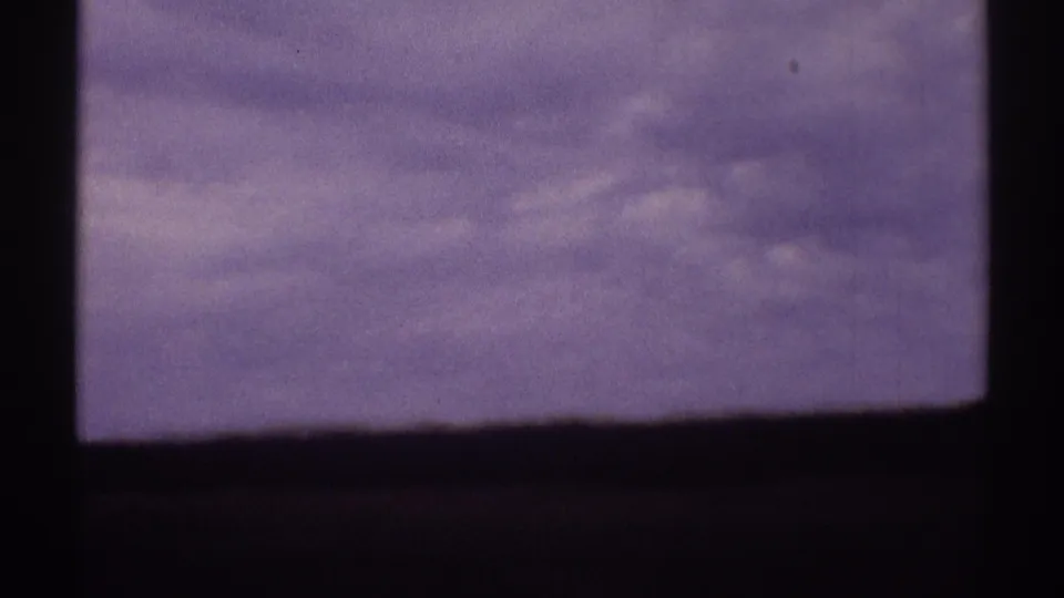 Overcast late-1960s 8mm roadside view with a low horizon, dark window framing, and purple-gray film color.