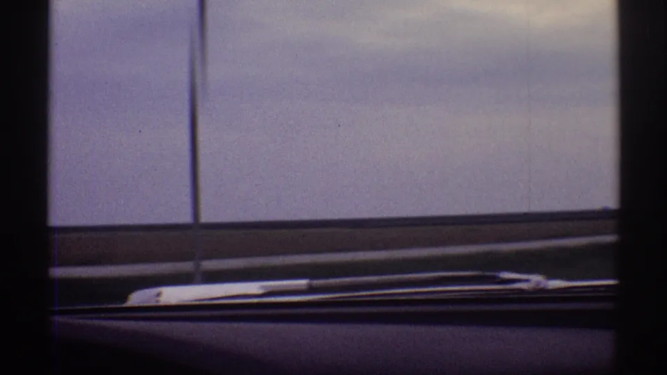 In-car 8mm view across a flat Illinois road landscape, with windshield posts, wiper blade, dashboard, and muted sky.