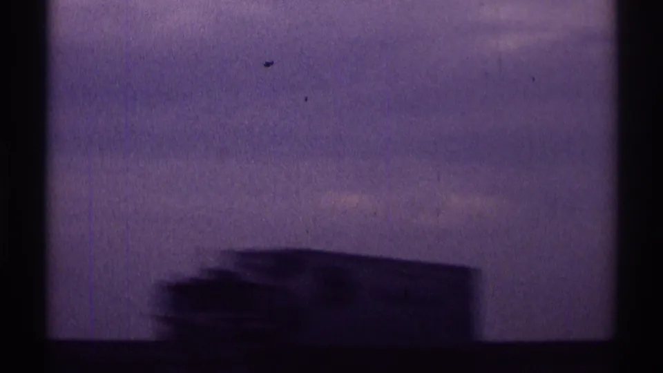 A blurred low roadside or vehicle-side form passes beneath a cloudy, purple-tinted sky in the moving car footage.