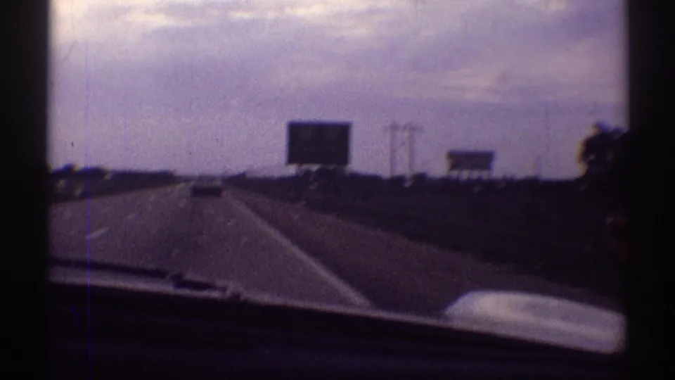 Windshield view of a highway with distant traffic, roadside signboards, power poles, and cloudy sky in 1969 Illinois home-movie footage.