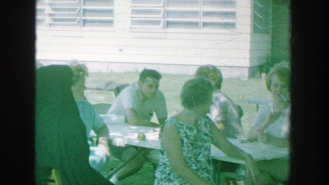 1964 Michigan home-movie frame showing adults gathered around a white outdoor table, with lawn chairs, grass, and louvered windows behind them.