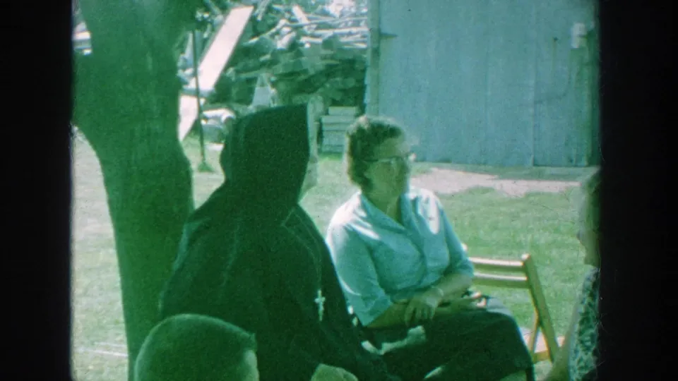 1964 Michigan home-movie frame showing adults in a backyard, including a seated woman with glasses and a figure in a dark habit near a small outbuilding.