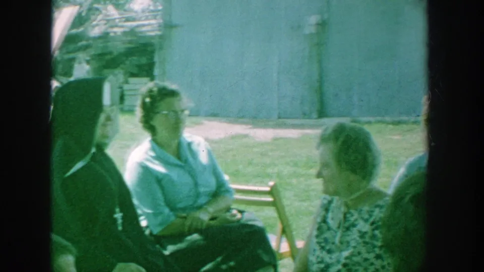 1964 Michigan backyard scene with two women in conversation on the lawn and a dark-clad religious figure partly visible nearby.