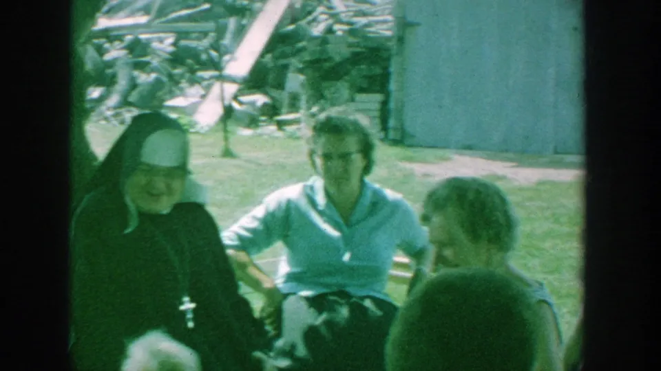 The nun remains seated with older women in a grassy Michigan yard, with a wooden structure and scattered debris behind them.