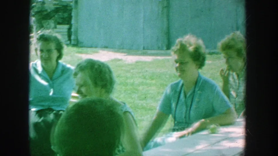 Older women sit around a picnic table in the yard, with one woman turned toward others during the outdoor gathering.