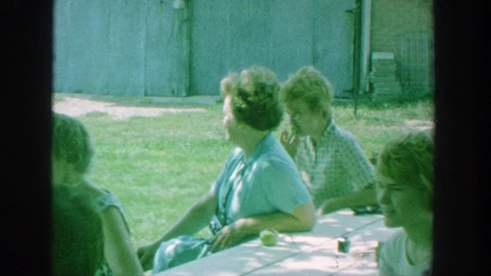 Women seated at a picnic table face different directions in the grassy Michigan yard, with the wooden shed visible behind them.