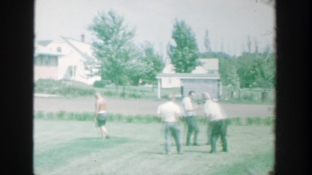 Several men stand and bend on a sunny lawn near houses and trees.