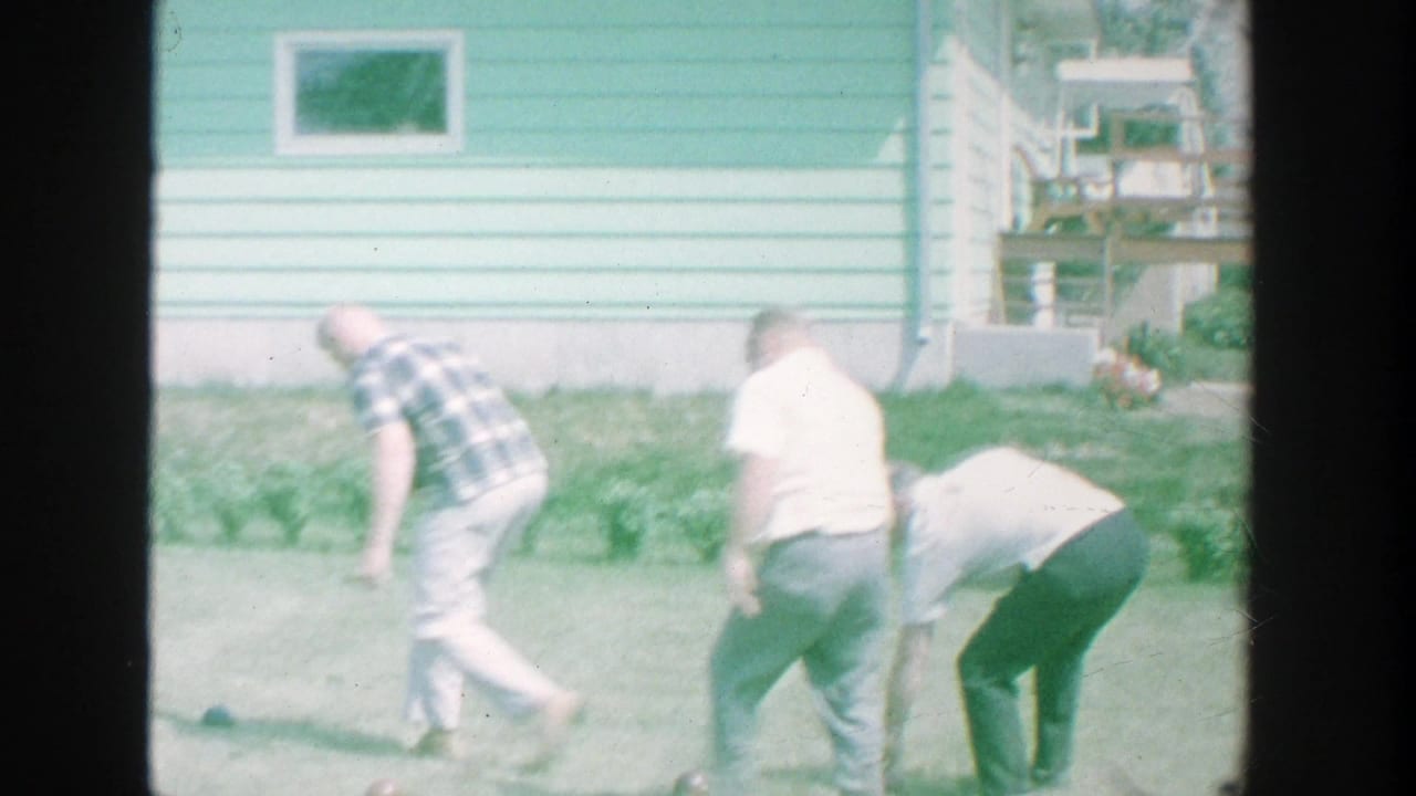 Men in casual clothing move across a Michigan backyard beside a light blue house, with small objects visible on the grass.