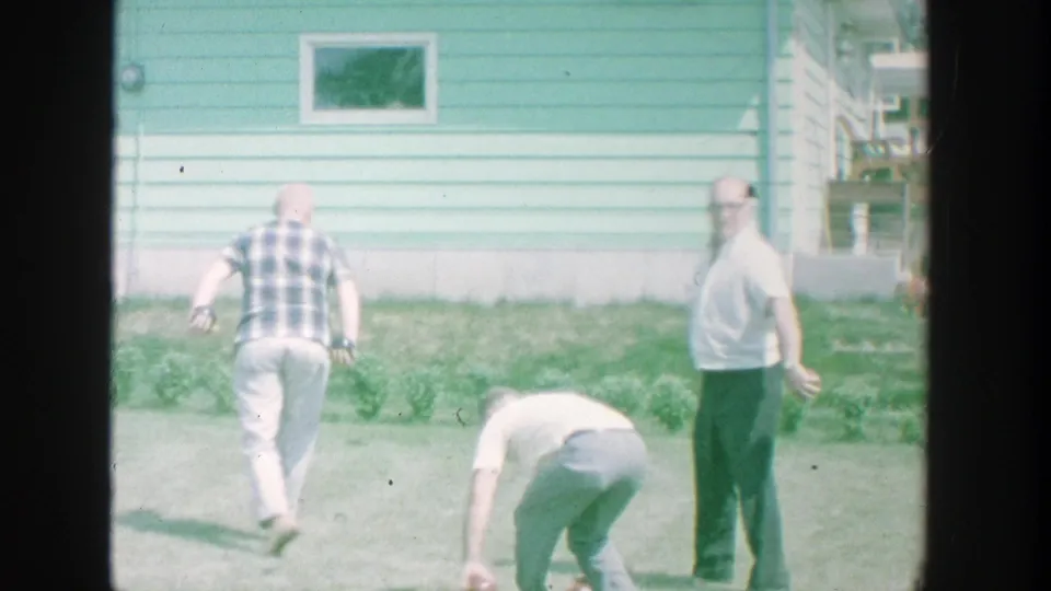 A 1964 Michigan backyard frame shows men near a light blue house, one bending to the grass while another looks toward the camera.