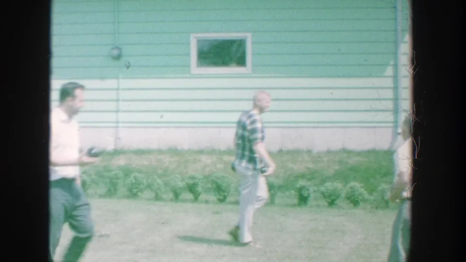 Men move through a sunny Michigan backyard in front of a light blue house, with one figure partly cropped at the frame edge.