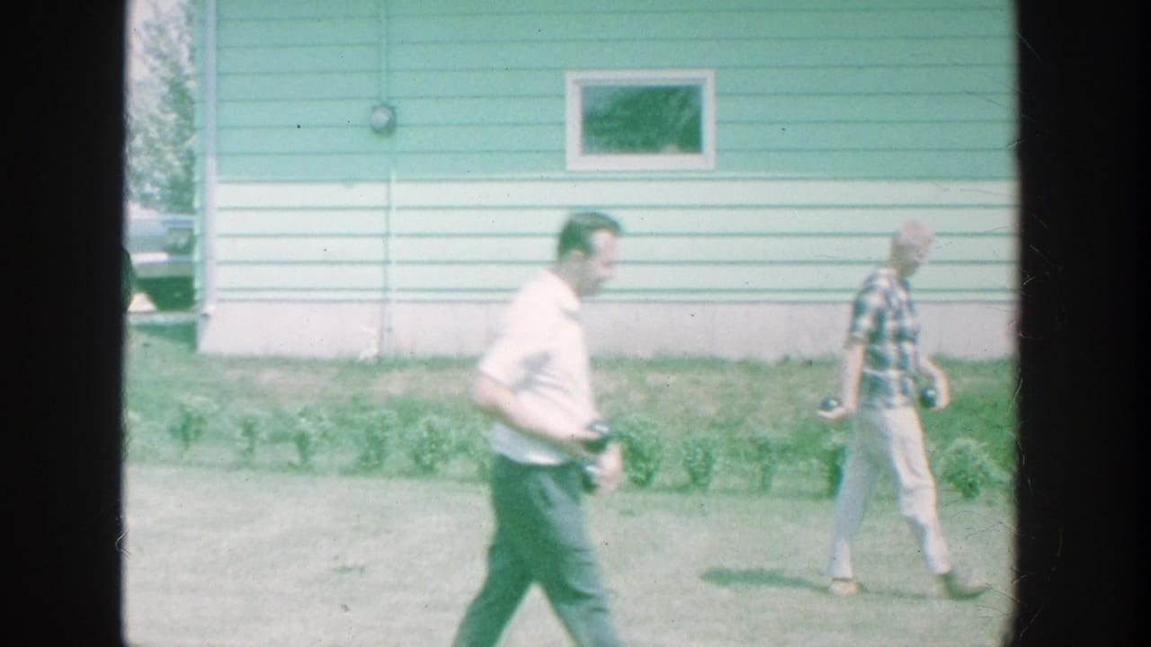 In supported 1964 Michigan footage, two men cross a lawn beside a pale green house, with garden plants along the wall.