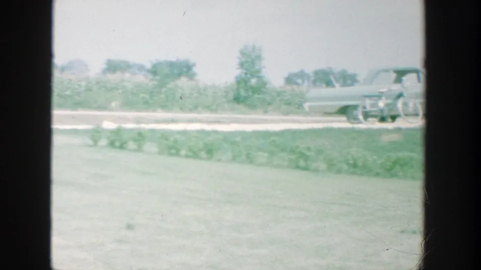 The frame shows a soft, green-tinted view of a lawn, garden row, and parked 1960s sedan near a rural-edge residence.