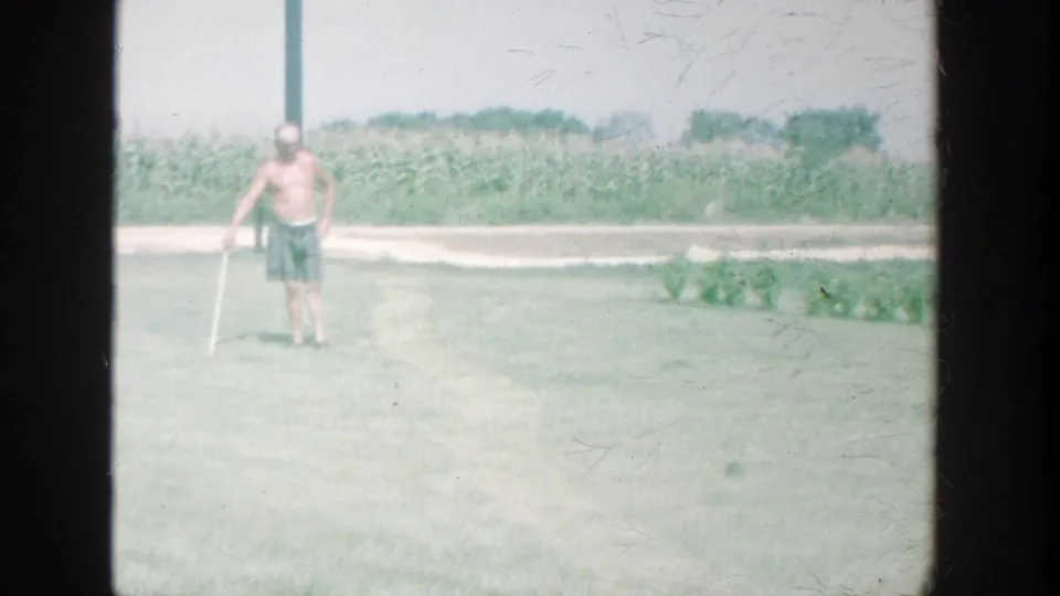 A shirtless adult man stands on a Michigan lawn holding a cane, with a cornfield and road edge visible behind him.
