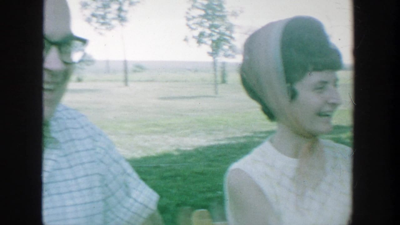 1964 Michigan 8mm frame showing a man in glasses and a woman in a white sleeveless dress seated or standing on grass.