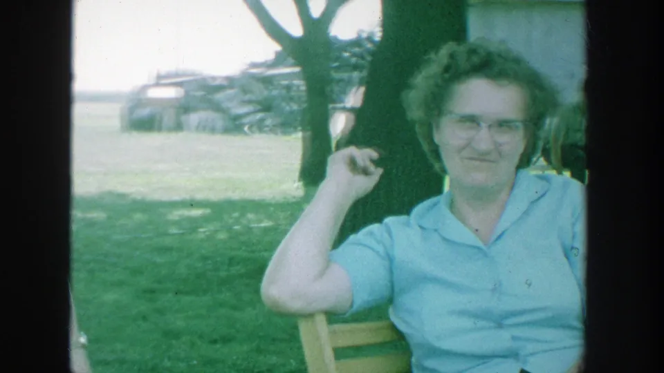 1964 Michigan 8mm frame showing a woman in glasses and a light blue collared shirt seated outdoors near a tree.