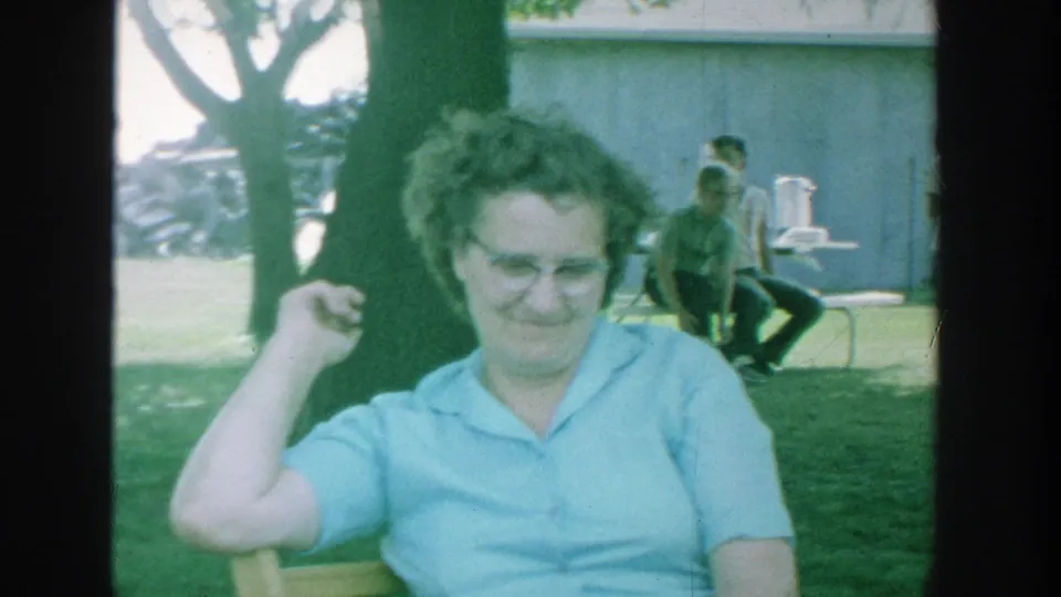 1964 Michigan home-movie frame with a woman in glasses in the foreground and seated adults in the background near a building.