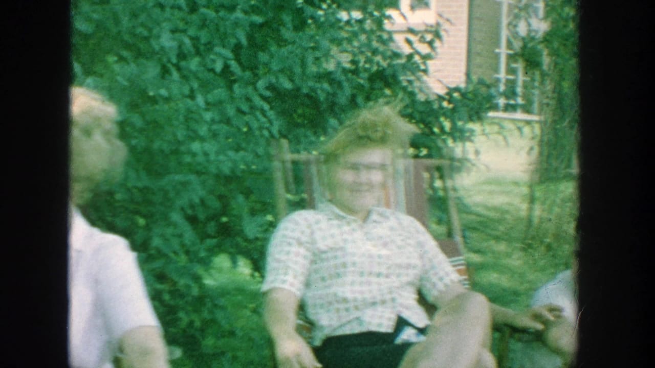 1964 Michigan home-movie frame of adults relaxing in a backyard, including a smiling woman in a patterned blouse near a brick house.