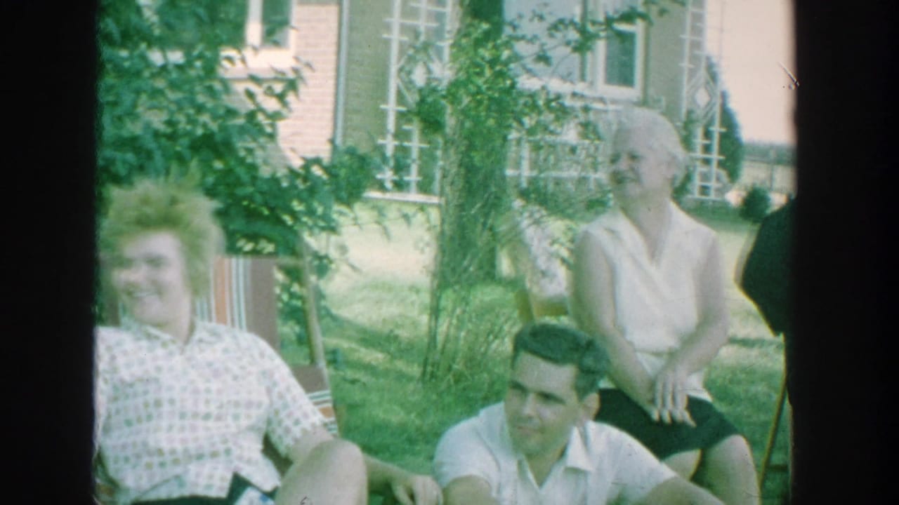 1964 Michigan backyard frame with seated adults, a woman in a sleeveless blouse, and a man on the grass near a brick house.