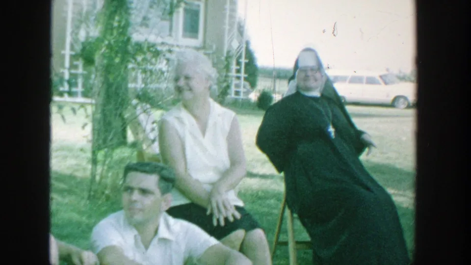 1964 Michigan home-movie frame showing a nun in a dark habit seated in a backyard with other adults nearby.