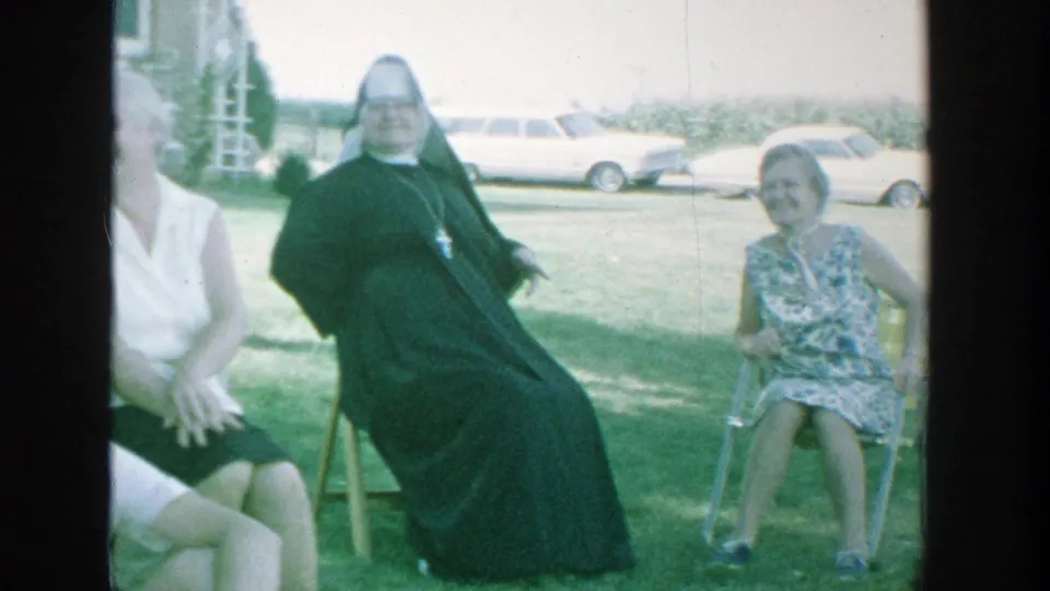 1964 Michigan backyard frame with a seated nun, a young woman in a patterned dress, and a light-colored station wagon behind them.