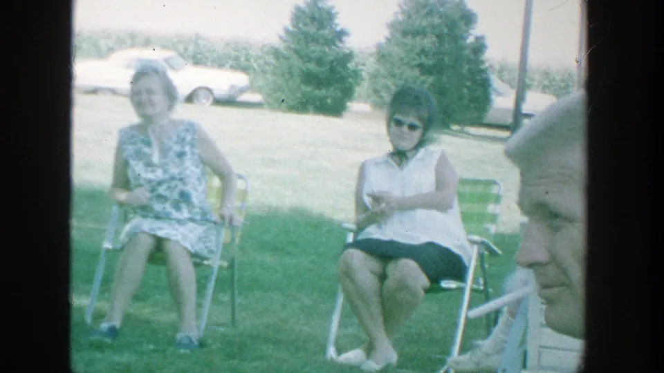 1964 Michigan park scene with two seated women, one in a floral dress and one wearing sunglasses, with classic cars in the background.