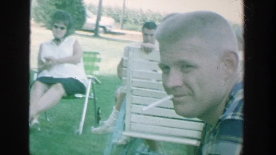 1964 Michigan home-movie frame with seated adults in a park, including a woman in sunglasses and a smiling bald man near the camera.