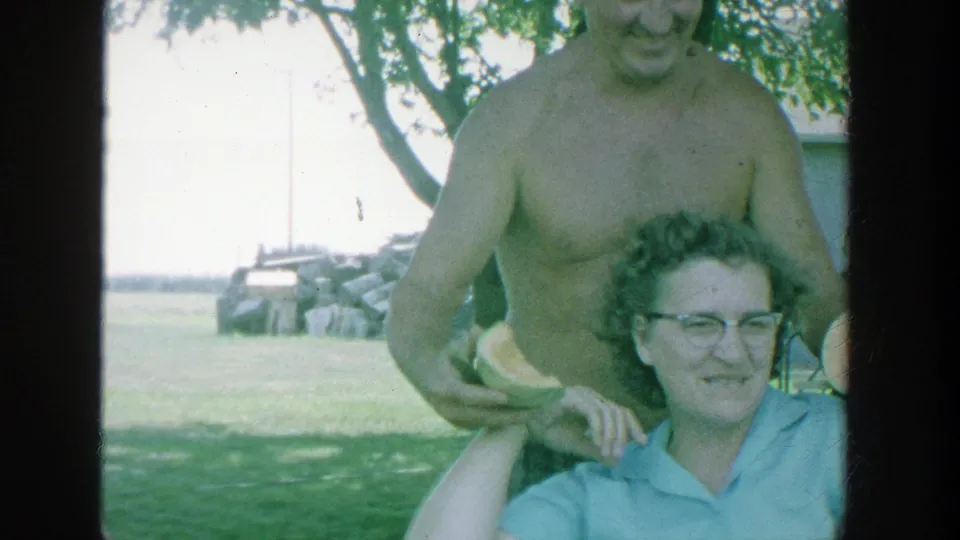 1964 Michigan park gathering frame showing a shirtless man handing watermelon to a smiling seated woman near shade and open lawn.