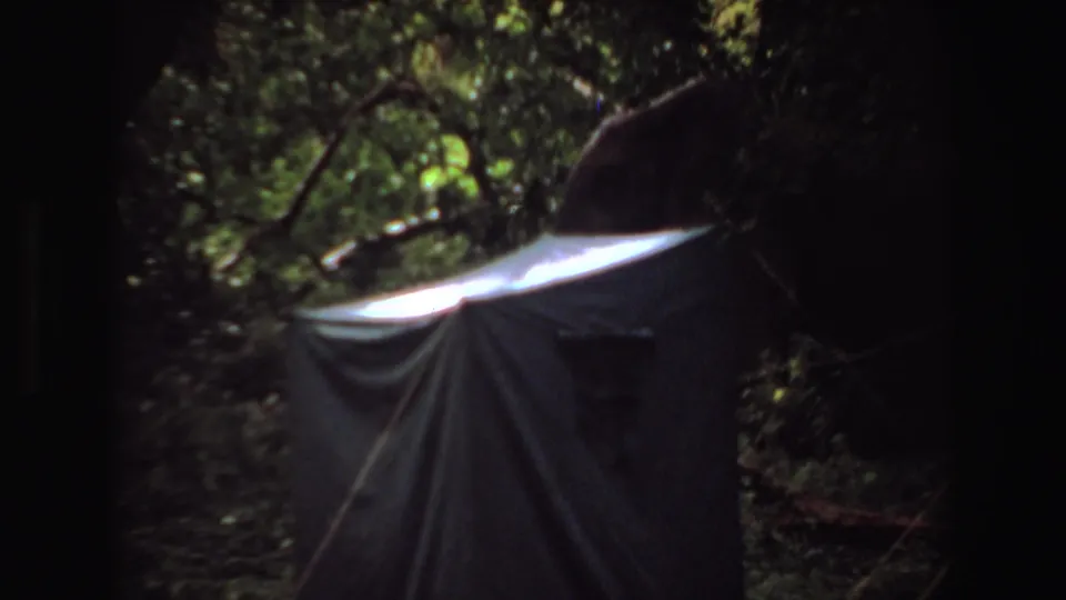 1969 Angola frame showing a grey tent or tarp structure among trees, with deep shadow around the frame and sunlit greenery above.