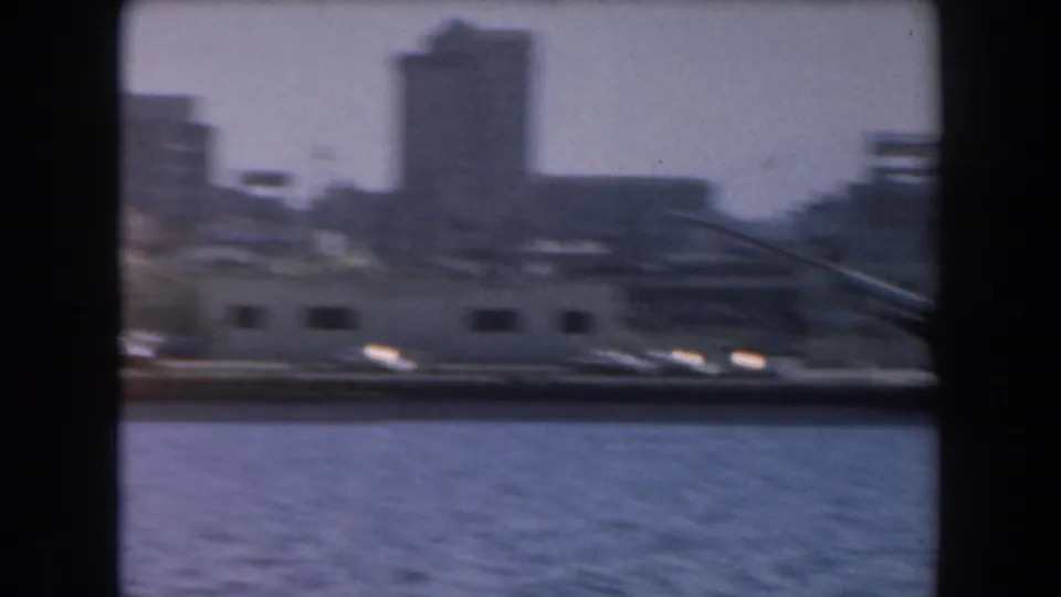 1961 California 8mm frame showing water in the foreground, low harbor buildings, and a blurred urban skyline beyond.