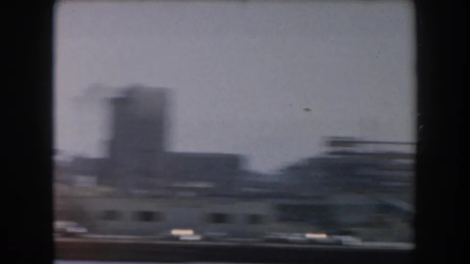 Motion-blurred 1961 California waterfront frame with open water, harbor structures, and a tall city building behind them.
