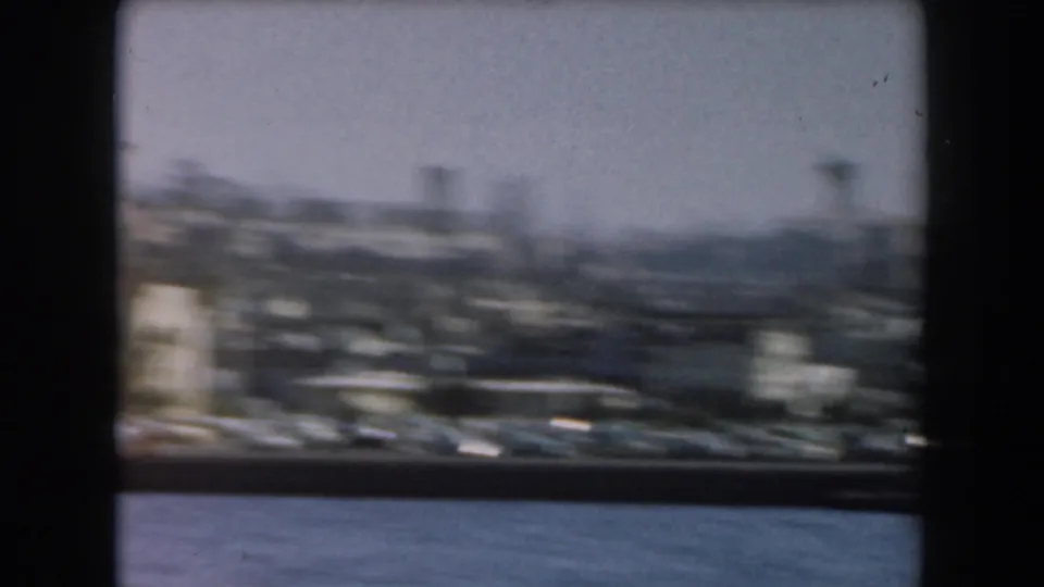 1961 California 8mm frame showing a hazy harbor area with boats, water in the foreground, and distant urban structures.